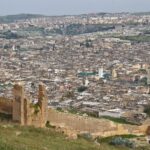 Panoramic view over the dense rooftops, minarets and green-tiled roofs of Fès el-Bali, seen from the ruined walls near the Merinid Tombs