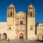 The twin bell towers and carved stone facade of the Church of Santo Domingo de Guzman in Oaxaca under a deep blue sky, with visitors walking on the plaza