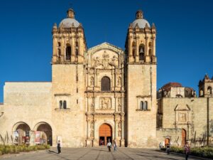 The twin bell towers and carved stone facade of the Church of Santo Domingo de Guzman in Oaxaca under a deep blue sky, with visitors walking on the plaza