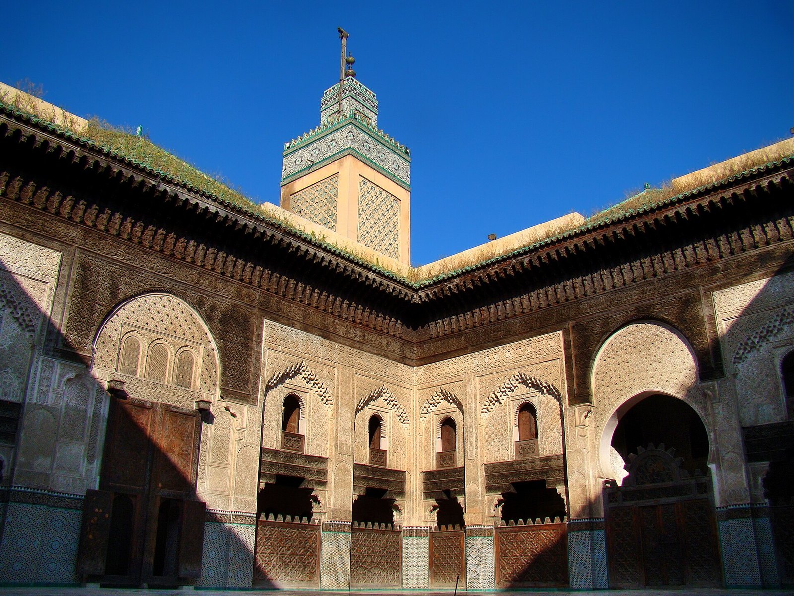 Courtyard of the Bou Inania Madrasa in Fez with carved stucco walls, cedar wood screens and the green-tiled minaret against a blue sky